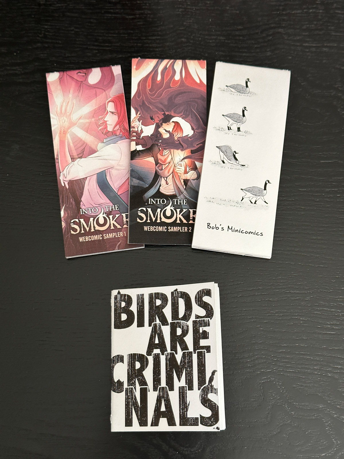 A photo of four mini zines. The three vertical zines are Into the Smoke Webcomic Sampler 1, Into the Smoke Webcomic Sampler 2, and Bob's Minicomics. The fourth square zine is Birds are Criminals.